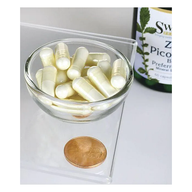 A small glass bowl of white capsules sits on a clear surface beside a penny for scale. Part of a Swanson supplement bottle labeled in green and yellow is visible in the background, suggesting its Zinc Picolinate 22 mg content, known for supporting the immune system.