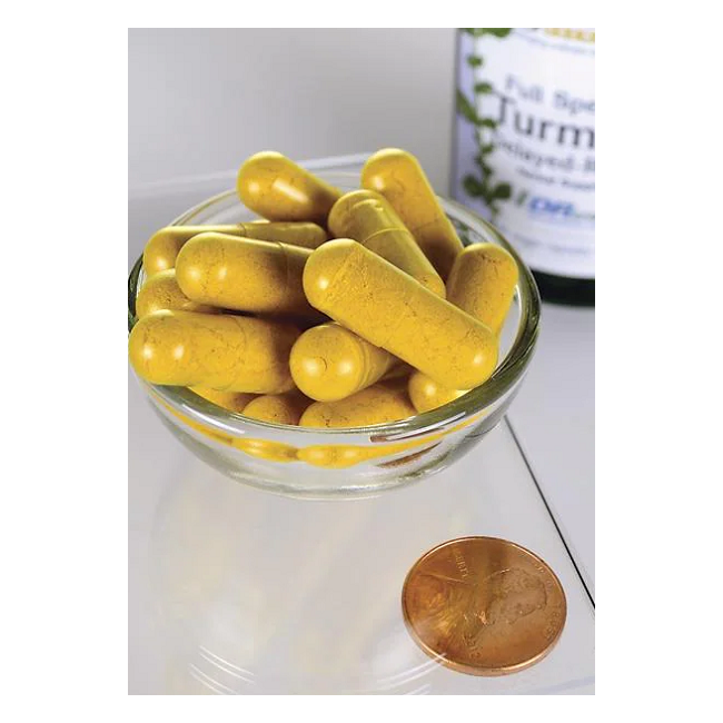 A glass bowl overflowing with yellow capsules sits next to a penny for scale, as a Swanson Turmeric 750 mg 60 Veggie EMBO Caps AP bottle is visible in the background.