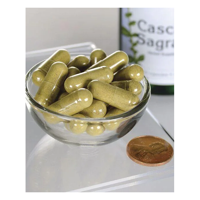 A small glass bowl of green capsules sits next to a penny for scale, offering gentle digestive support. A blurred bottle in the background is labeled "Swanson Cascara Sagrada 450 mg 100 Capsules.