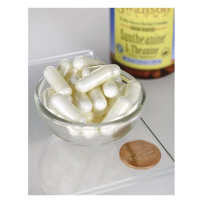 A glass bowl of Swanson Suntheanine L-Theanine 100 mg veggie capsules is placed beside a penny for scale, suggesting their compact power. In the background, a partially visible bottle indicates these capsules promote relaxation and contain L-theanine, found in green tea.