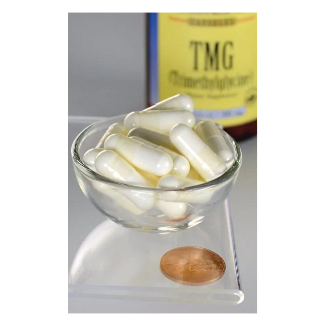 A glass bowl filled with white capsules sits next to a copper coin, with a Swanson TMG Trimethylglycine bottle suggesting its role in promoting liver health.