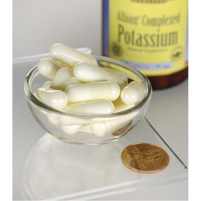 A glass bowl filled with Swanson Albion Chelated Potassium Glycinate 99 mg 90 capsules next to a container of potassium supplement and a penny for scale.